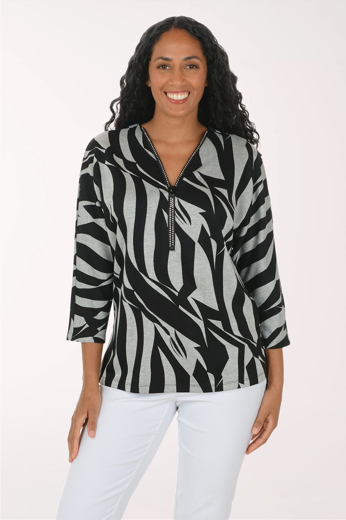 A woman modeling a black and gray abstract patterned top with a half-zip neckline, paired with white pants, against a plain light background.