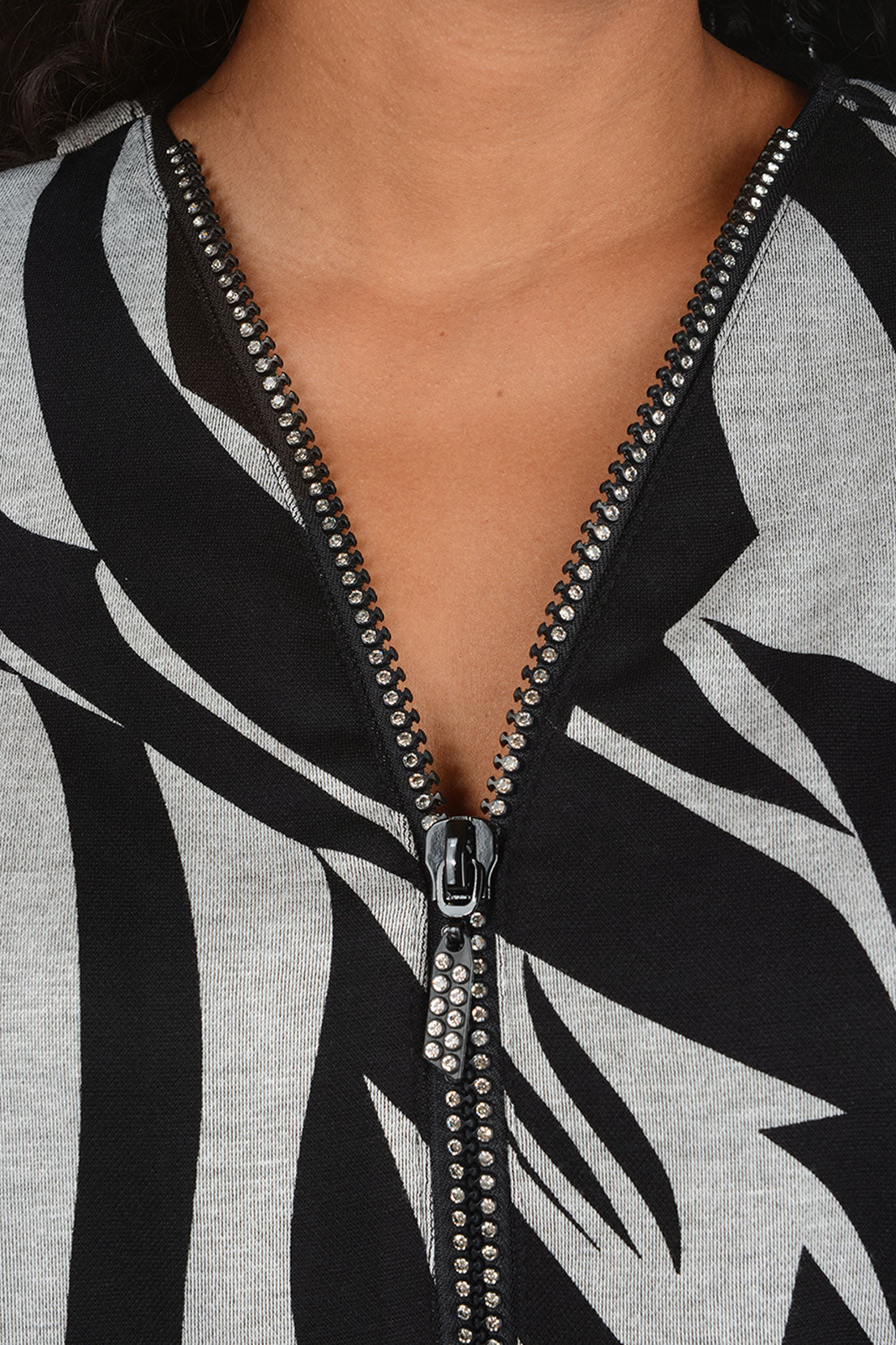 A woman modeling a black and gray abstract patterned top with a half-zip neckline, paired with white pants, against a plain light background.