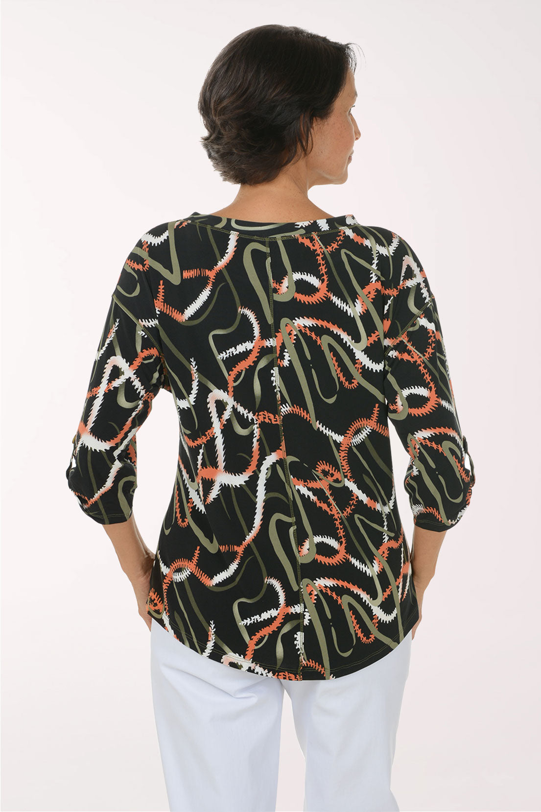 A smiling woman wearing a black three-quarter sleeve top with an abstract wavy pattern in white, orange, and olive green, paired with white pants, against a plain white studio background.