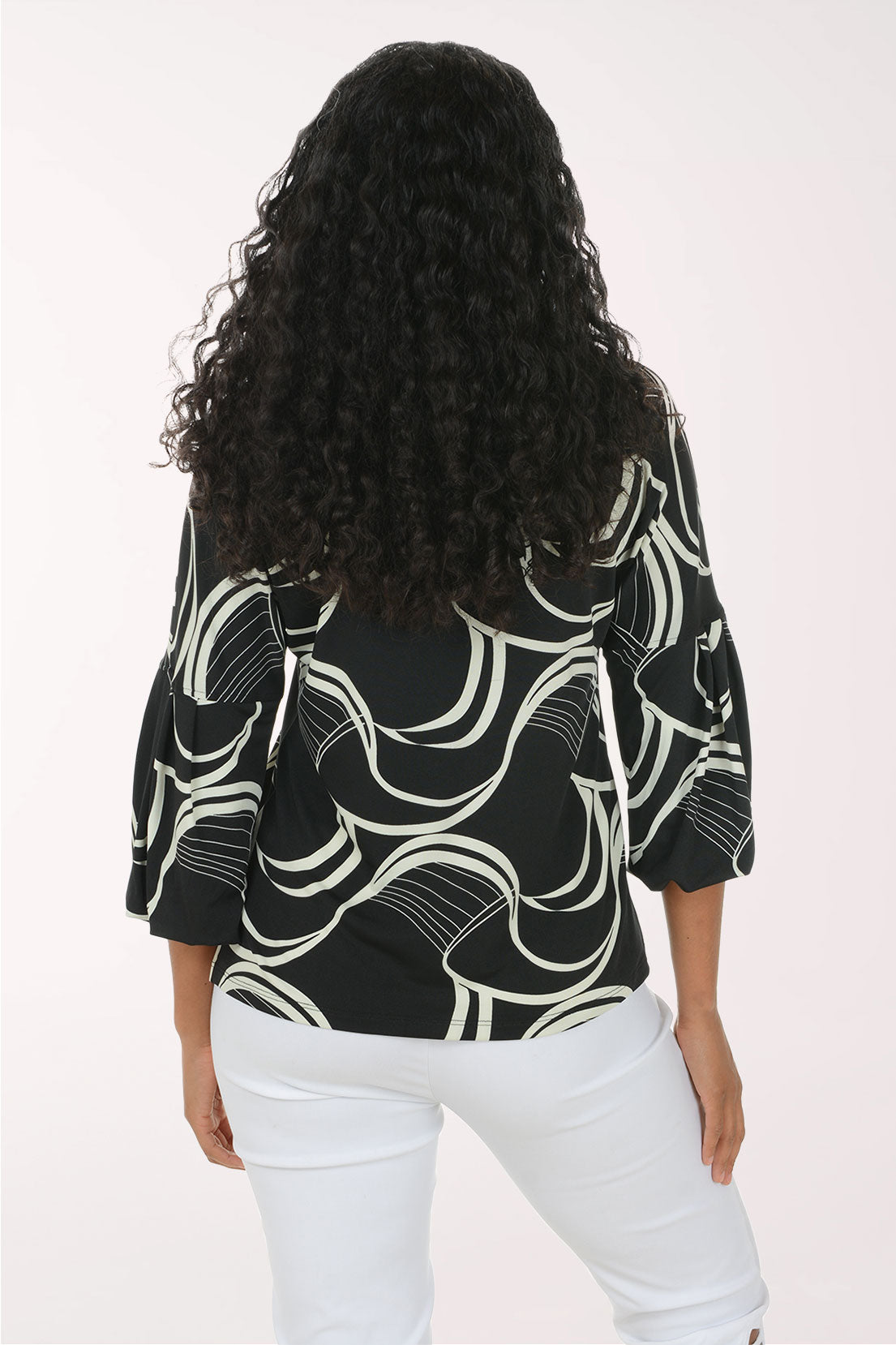 A woman viewed from behind wearing a black three-quarter sleeve blouse with a white abstract wavy line pattern, paired with white pants, against a plain light background.