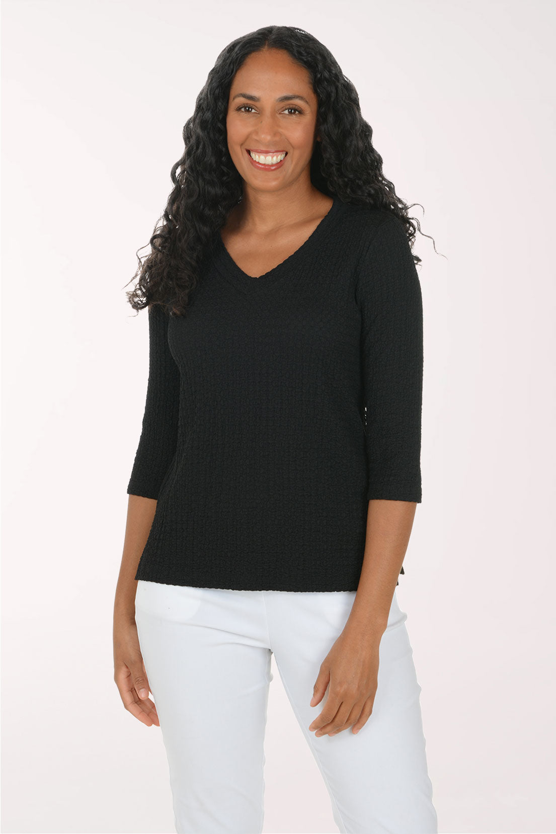 A woman with long, curly black hair wearing a black ribbed three-quarter sleeve V-neck top and white pants, standing against a plain light background.