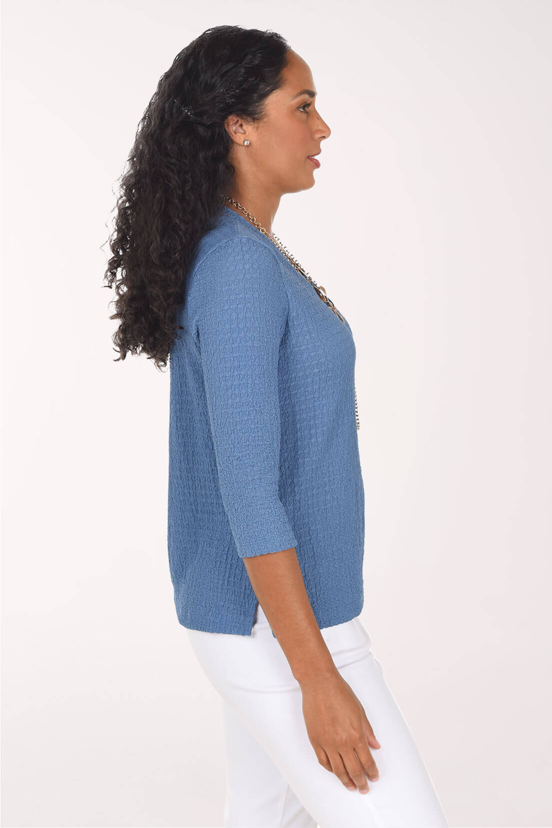 A side shot of a woman with long, curly black hair wearing a blue ribbed three-quarter sleeve V-neck top, a multi-strand chain necklace, and white pants, standing against a plain light background.