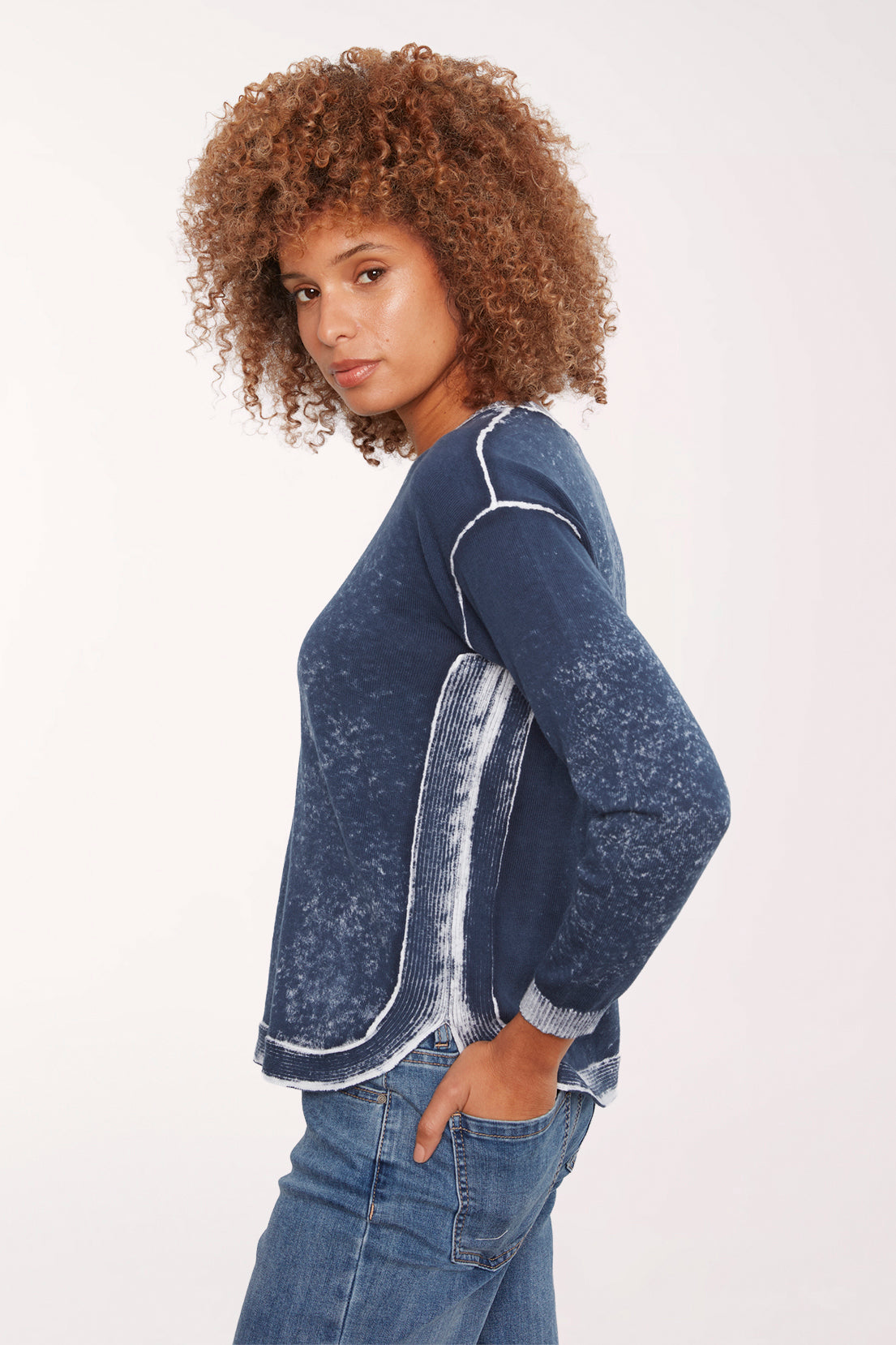 A woman with curly brown hair wearing a dark blue acid-wash long-sleeve top with white contrast stitching and blue jeans, photographed against a plain white studio background.