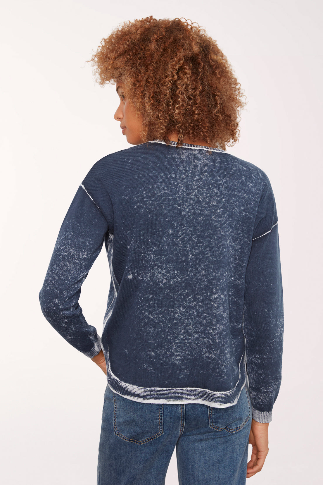 A woman with curly brown hair wearing a dark blue acid-wash long-sleeve top with white contrast stitching and blue jeans, photographed against a plain white studio background.
