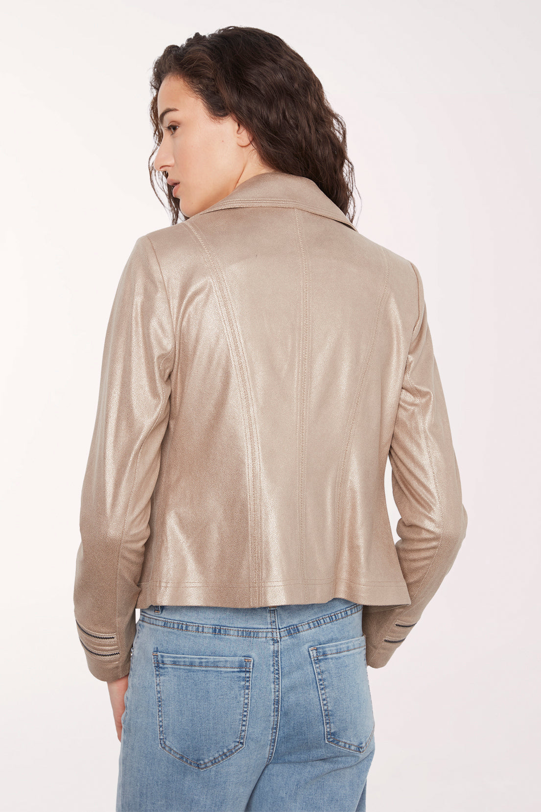 A woman shown from the back wearing a cropped, shiny champagne-beige faux leather jacket and light-wash blue jeans against a plain white studio background.
