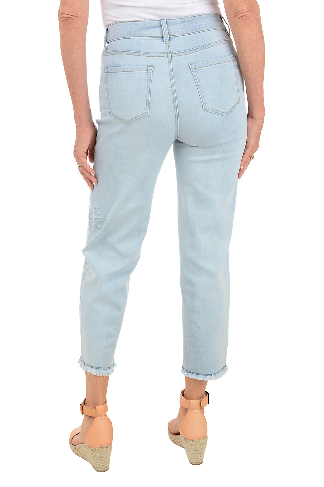 A pair of light blue denim ankle pants with embroidery on the left leg and frayed hemline, displayed on a model wearing tan sandals. Back shot.