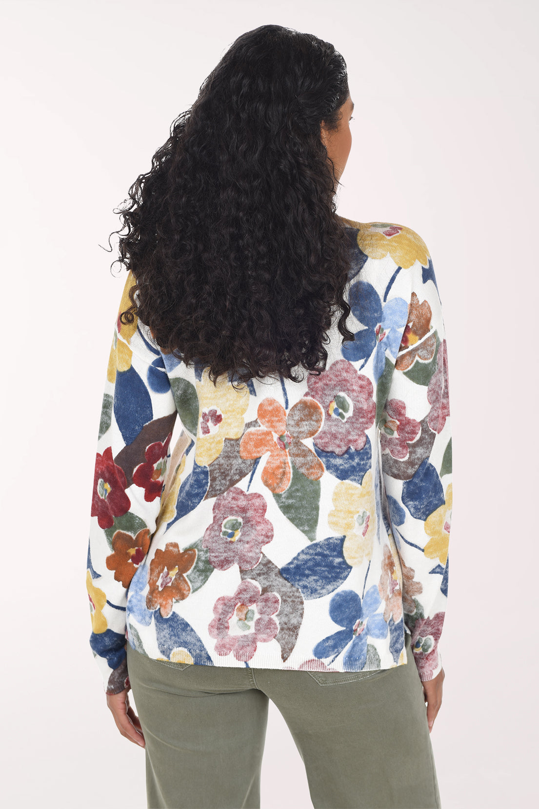 A woman wearing a white long-sleeve V-neck top with a colorful floral print in blue, red, yellow, and orange, paired with olive green pants, against a plain light background.