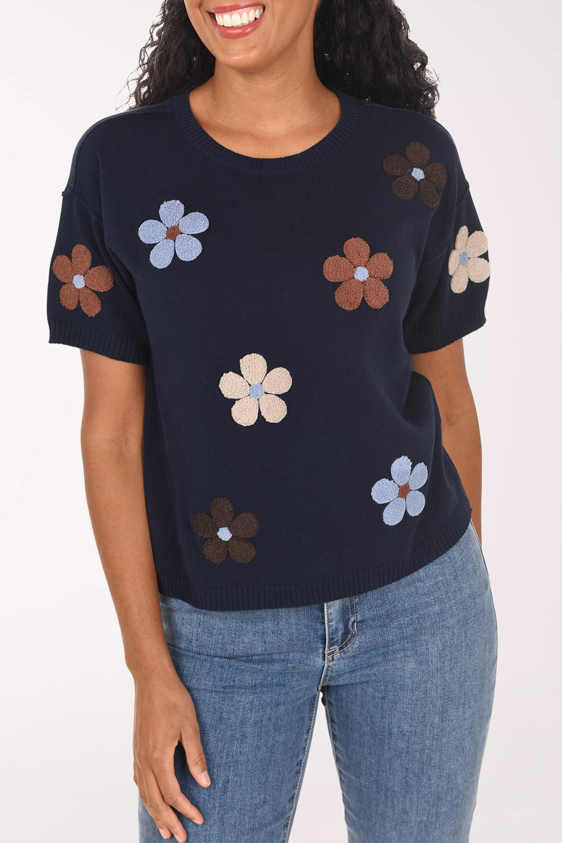 A woman modeling a navy blue short-sleeve top with embroidered flowers in brown, light blue, and cream, paired with blue jeans against a plain white background.