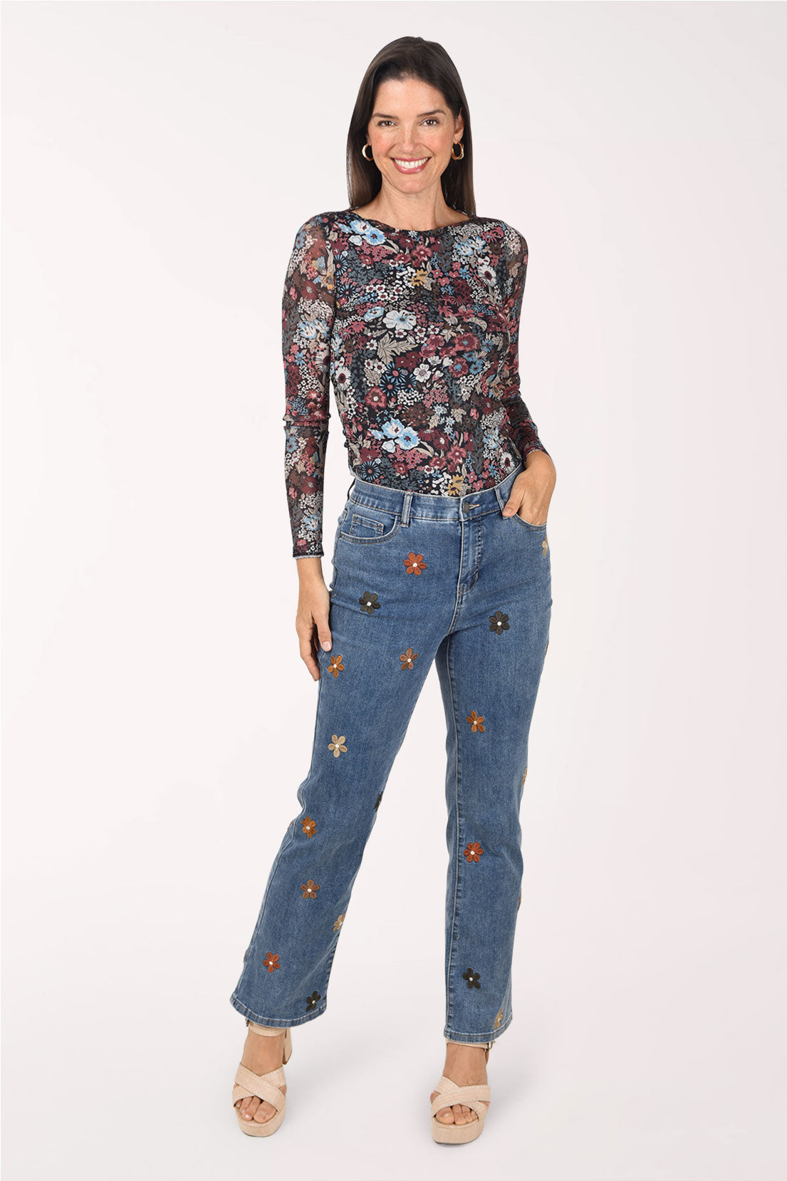 A woman wearing medium blue straight leg jeans with small embroidered brown and black flowers, styled with a dark floral print long sleeve top and heeled sandals.