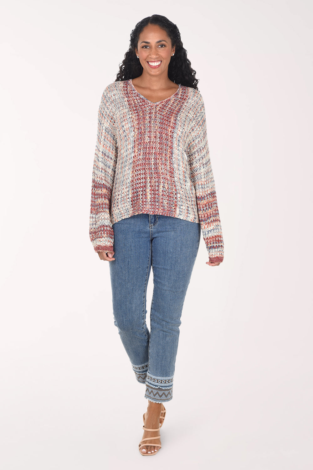 A woman modeling a long-sleeve knit top with a textured, multicolor pattern of red, blue, and cream, paired with blue jeans and beige heeled sandals, against a plain white studio background.