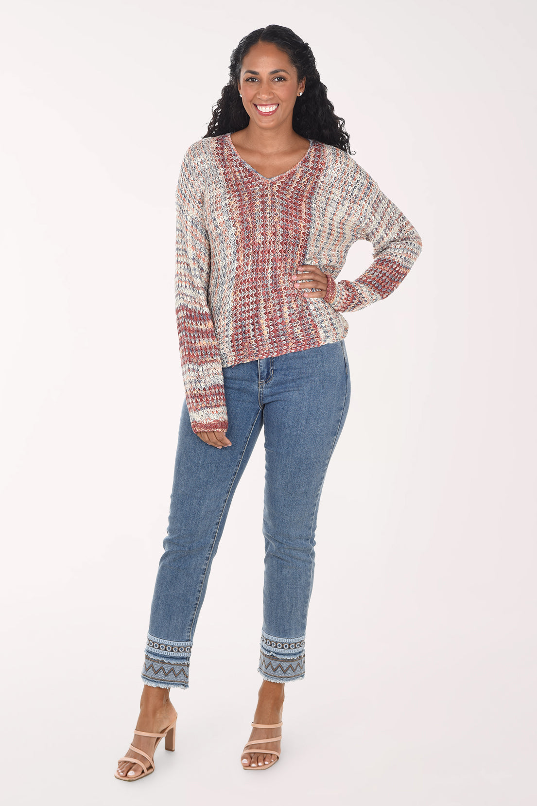 A woman modeling a long-sleeve knit top with a textured, multicolor pattern of red, blue, and cream, paired with blue jeans and beige heeled sandals, against a plain white studio background.