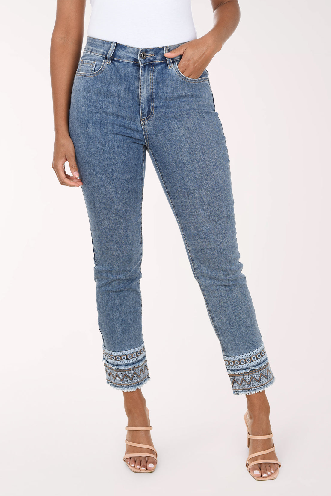 A model wearing a pair of medium-wash blue denim jeans with a geometric pattern trim at the rolled cuffs, paired with a white top and beige strappy heels, against a plain light background.