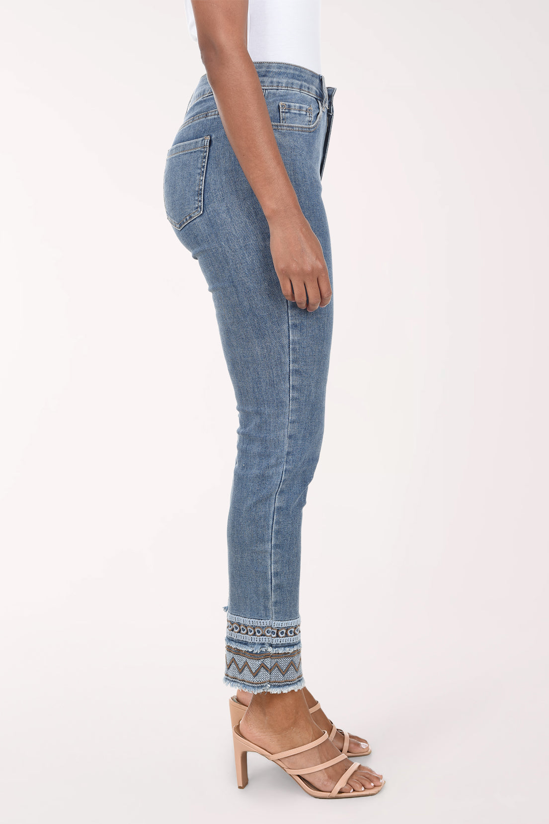 A model wearing a pair of medium-wash blue denim jeans with a geometric pattern trim at the rolled cuffs, paired with a white top and beige strappy heels, against a plain light background.