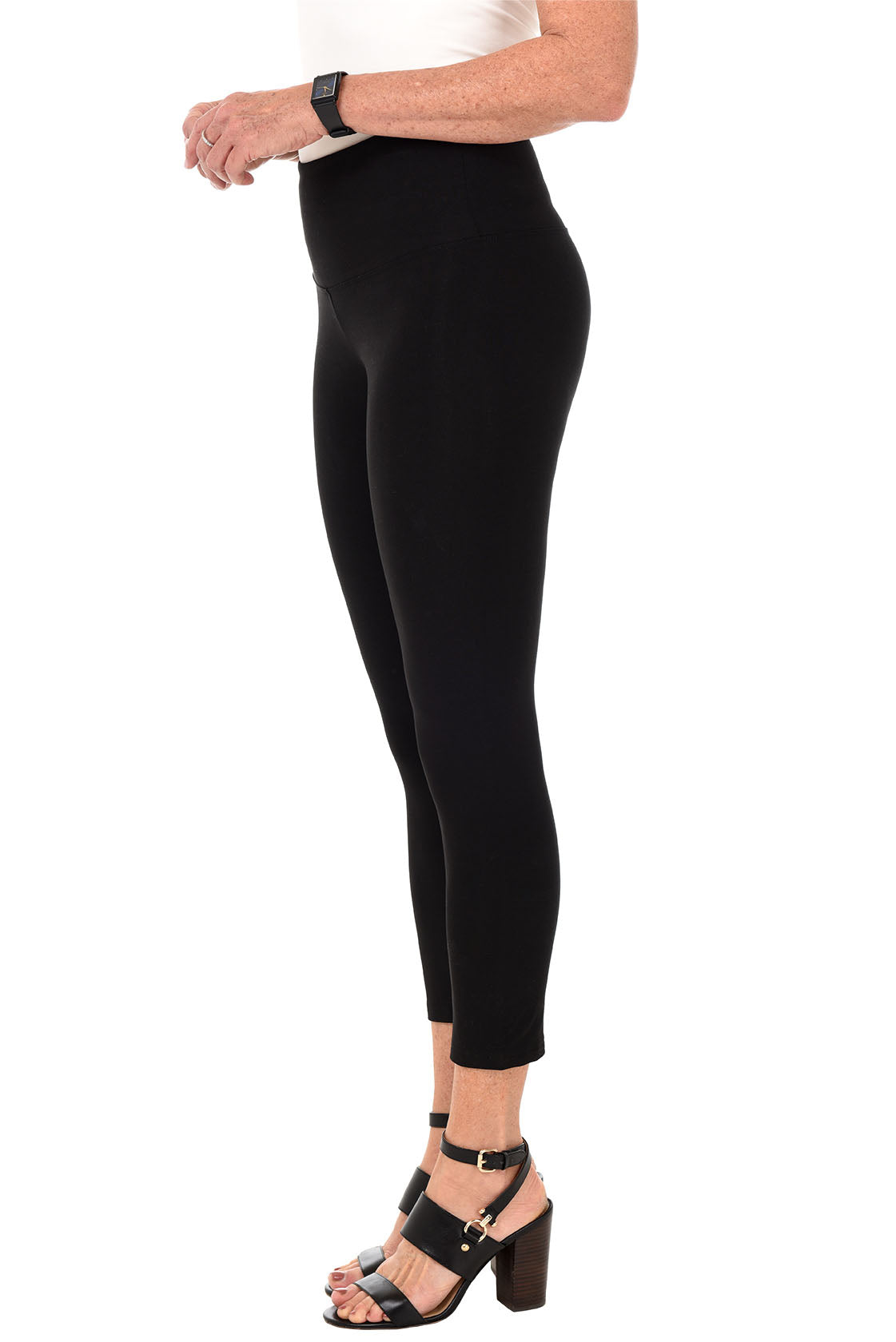 A side shot of a model wearing a pair of black pull-on cropped leggings with a wide elasticized waistband paired with black heels.