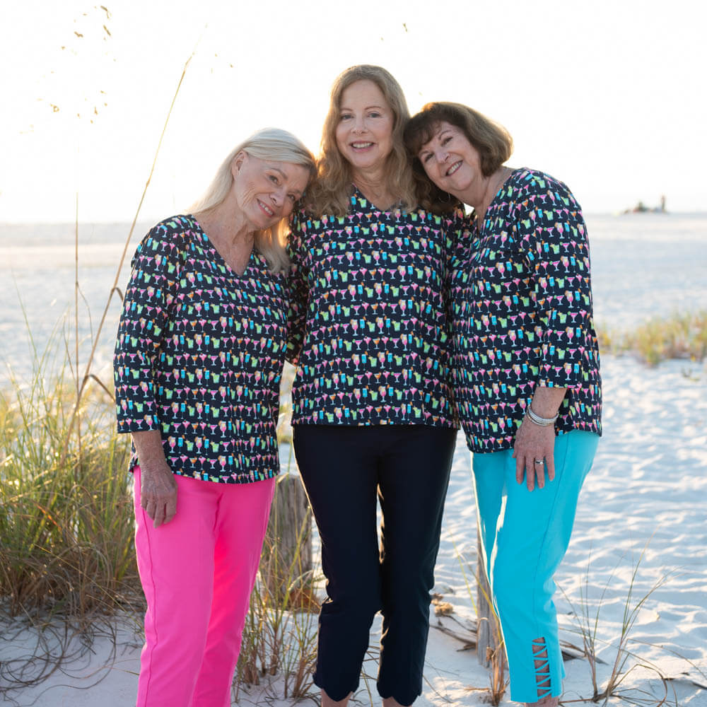 Three women wearing tropical drink print tops and colorful pants standing on a sandy beach.