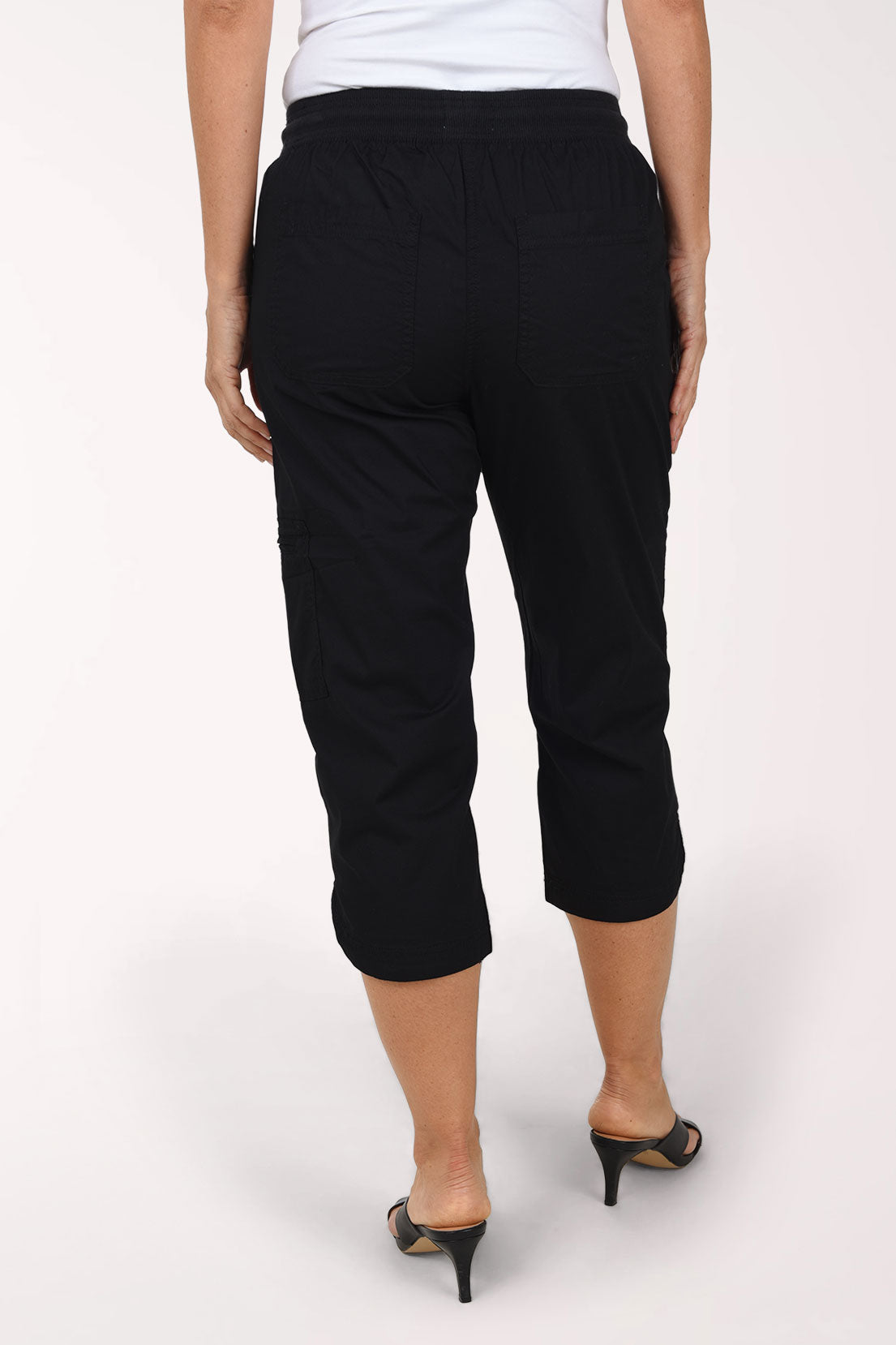 Back view of black capri pants with a smooth rib knit waistband, two back patch pockets and stitched seams down the leg, worn with a white top and black heeled sandals.