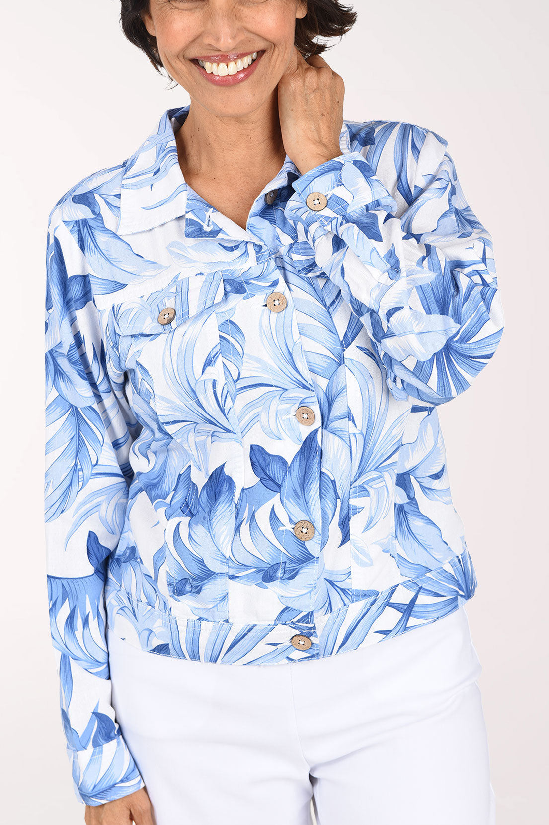 A woman wearing a blue and white tropical floral print button-up jacket and white pants, standing against a plain light background.