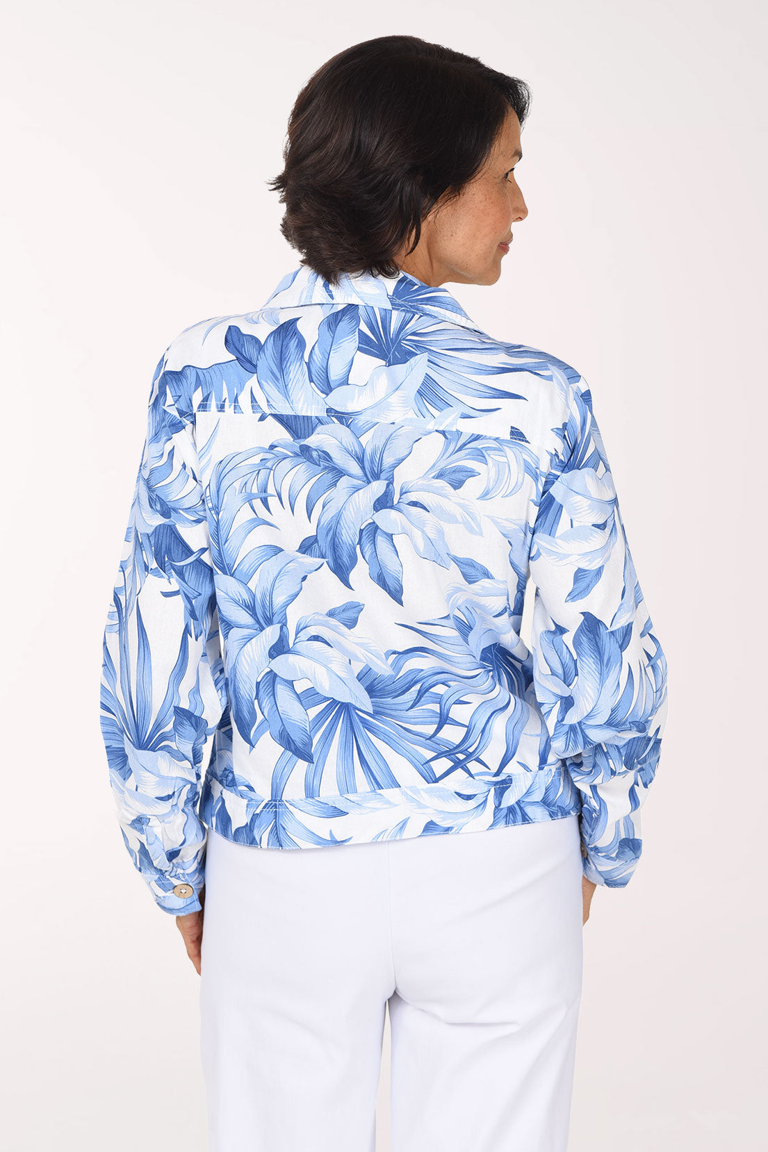 A woman wearing a blue and white tropical floral print button-up jacket and white pants, standing against a plain light background.