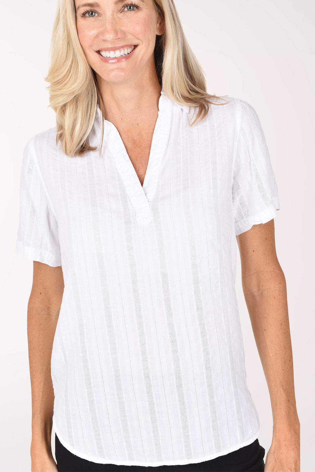 A smiling blonde woman wearing a white short-sleeve button-front blouse with subtle vertical texture and black pants, standing against a plain light background.