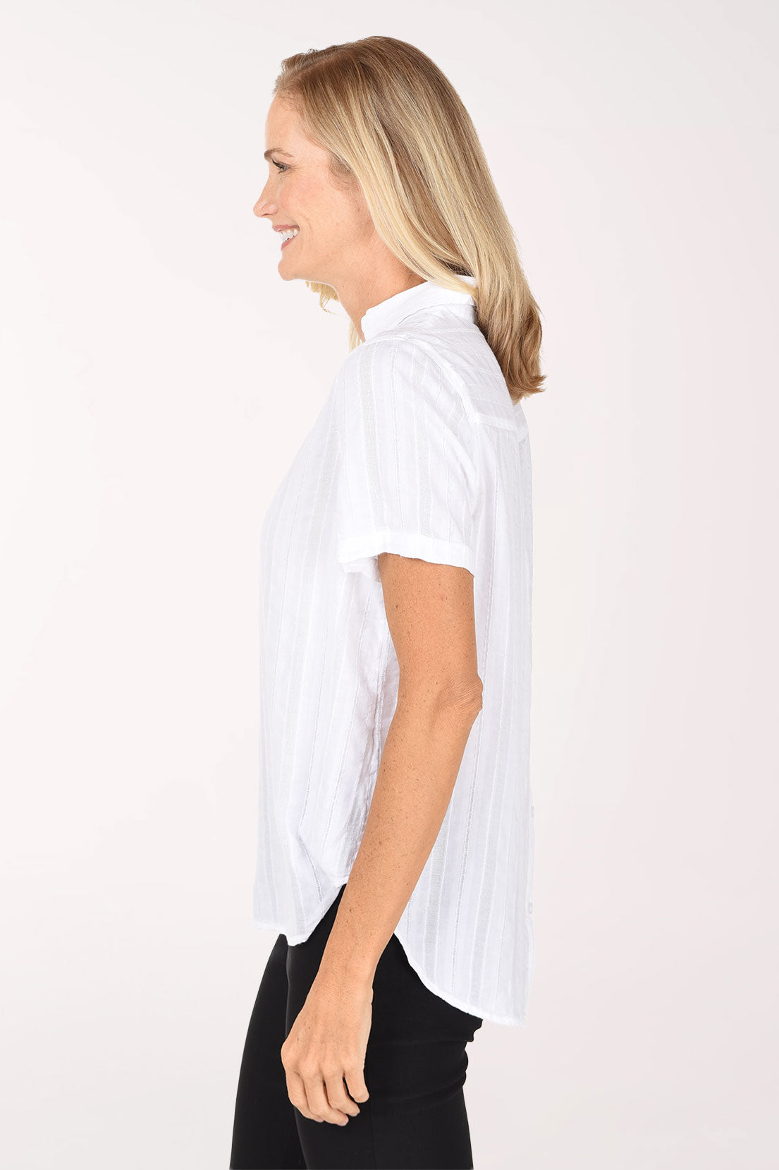 A smiling blonde woman wearing a white short-sleeve button-front blouse with subtle vertical texture and black pants, standing against a plain light background.