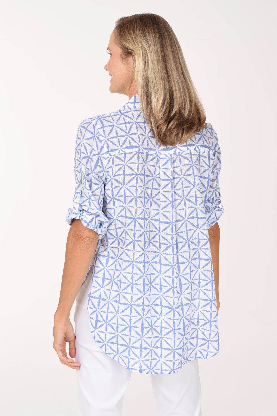 A smiling blonde woman wearing a light blue and white geometric patterned button-down shirt, posing against a solid off-white studio background.