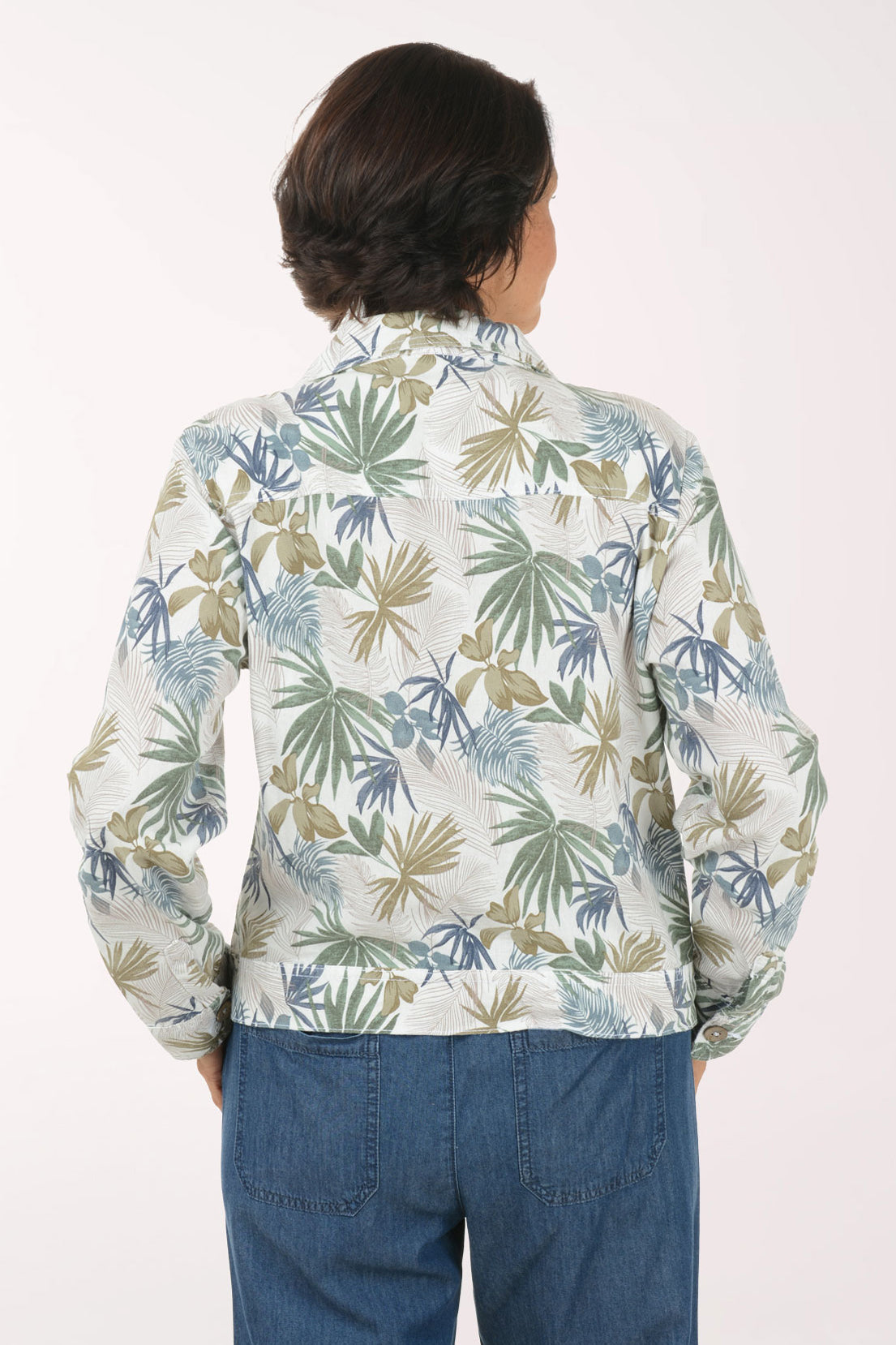 A woman wearing a long-sleeve button-up shirt with a blue, green, and beige palm leaf pattern, paired with blue jeans, posing against a solid white background.