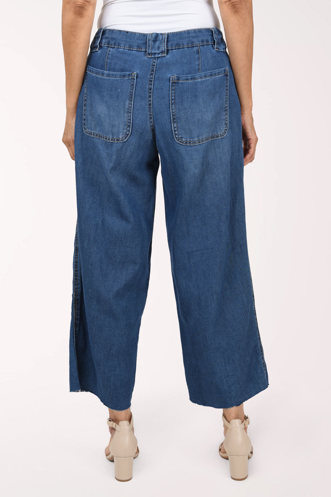 A model wearing a pair of wide-leg cropped blue denim jeans with a white top and beige heeled sandals, shown from the waist down against a plain white studio background.