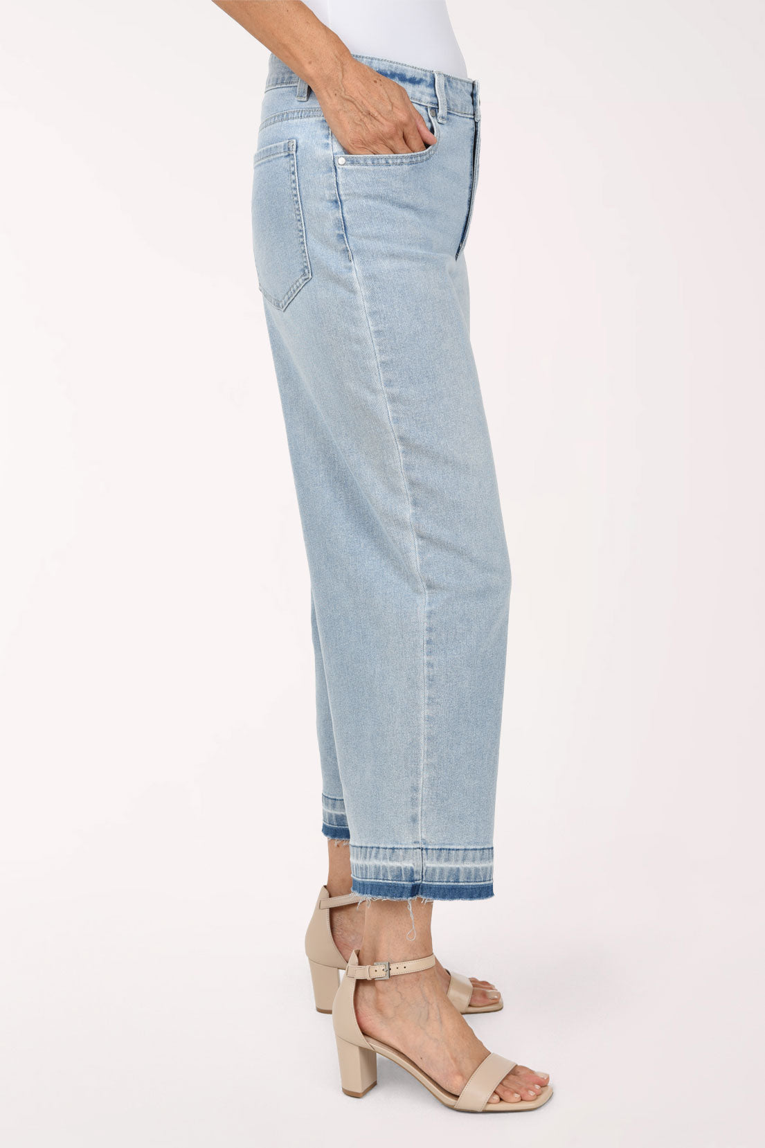 A person wearing light-wash cropped denim jeans with a straight-to-wide leg, shown from the waist down against a plain light background. The model has their hands in the pockets and is wearing beige heeled sandals.