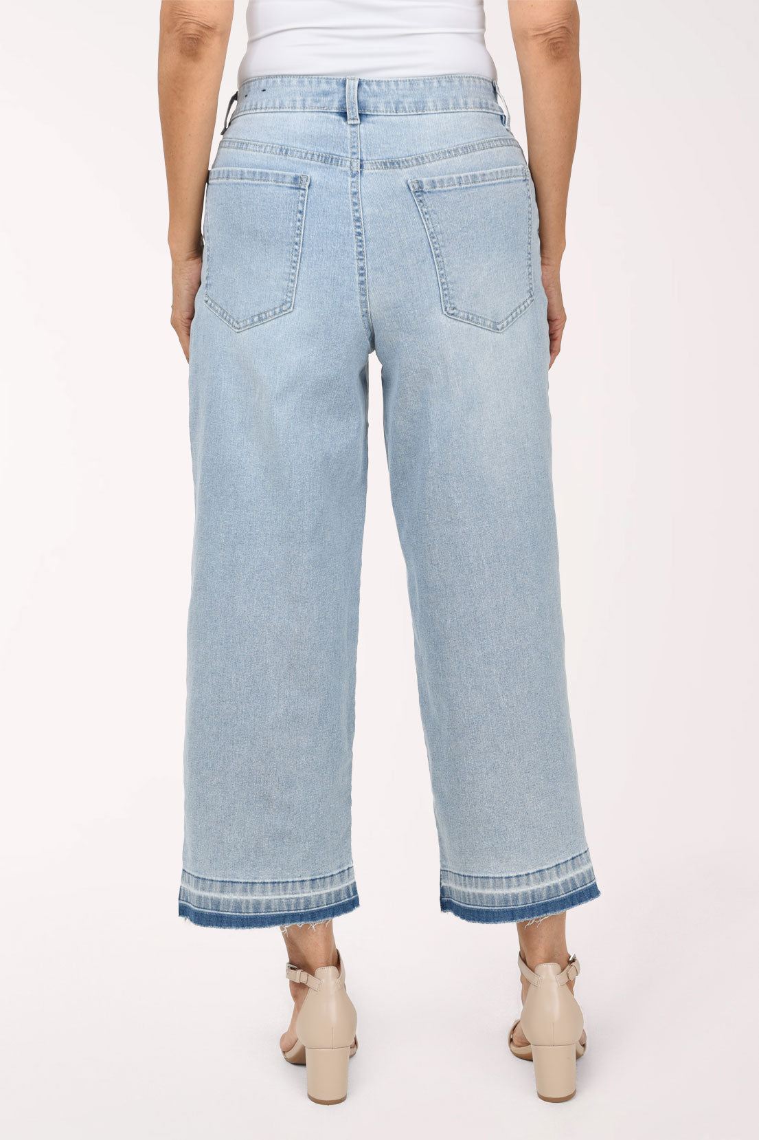 A person wearing light-wash cropped denim jeans with a straight-to-wide leg, shown from the waist down against a plain light background. The model has their hands in the pockets and is wearing beige heeled sandals.