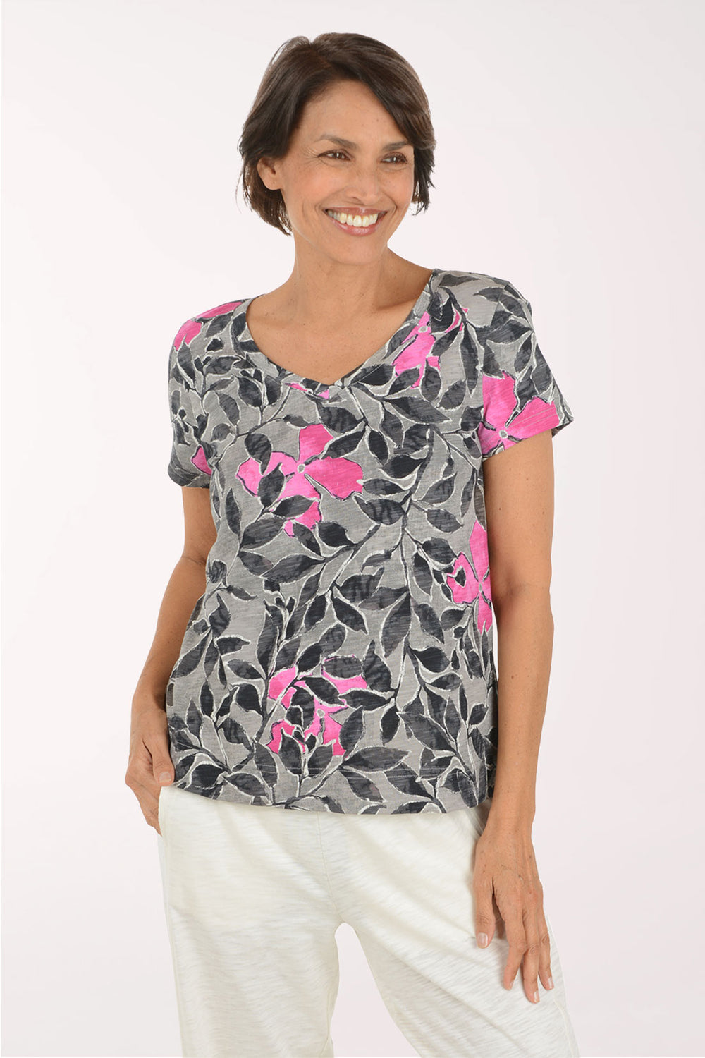 A woman wearing a gray short-sleeve V-neck top with a black leaf pattern and bright pink floral accents, paired with white pants, against a plain light background.