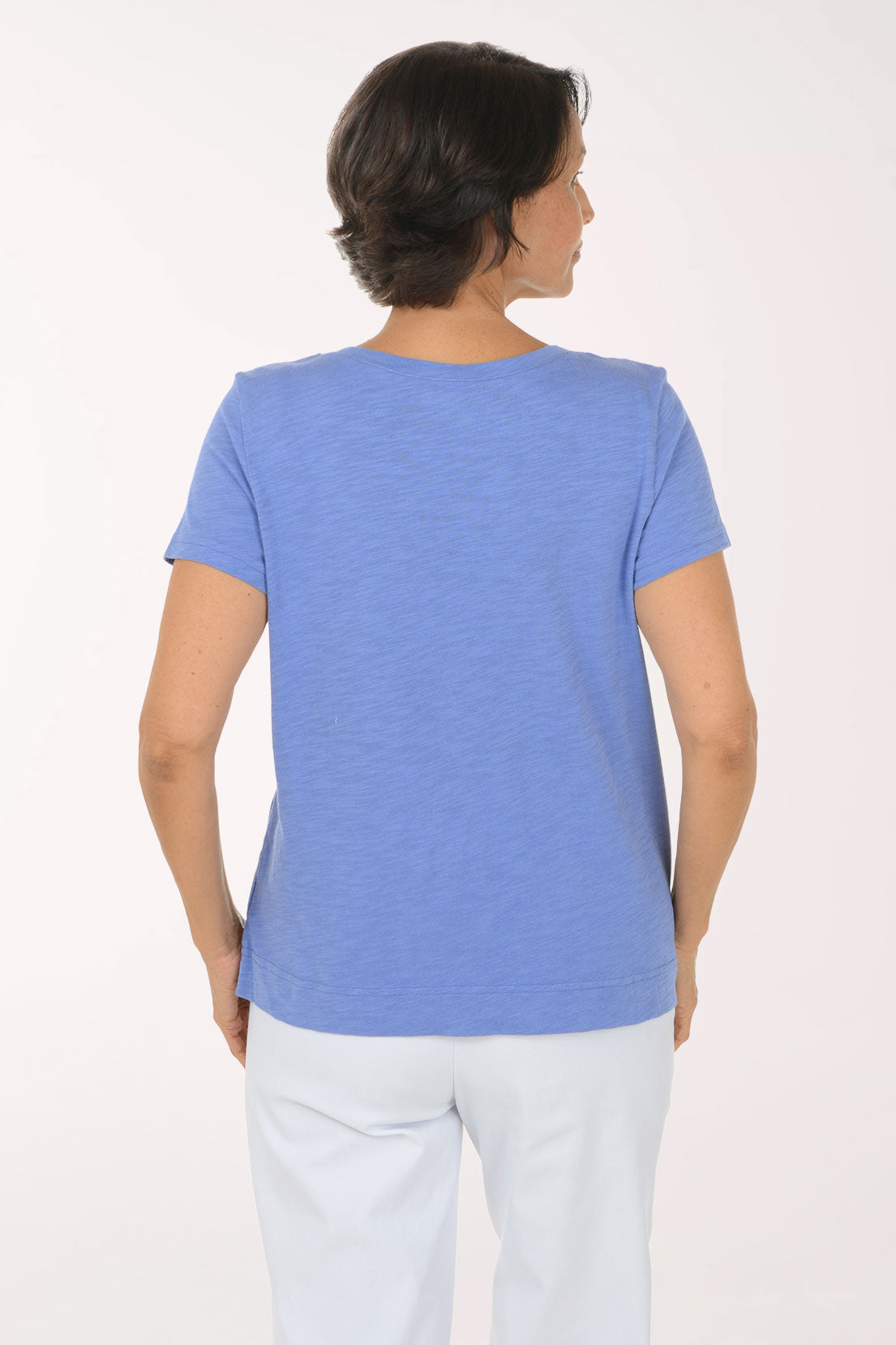 A smiling woman wearing a heathered periwinkle blue V-neck t-shirt with a white heart-shaped graphic on the front, paired with white pants, against a plain white studio background.