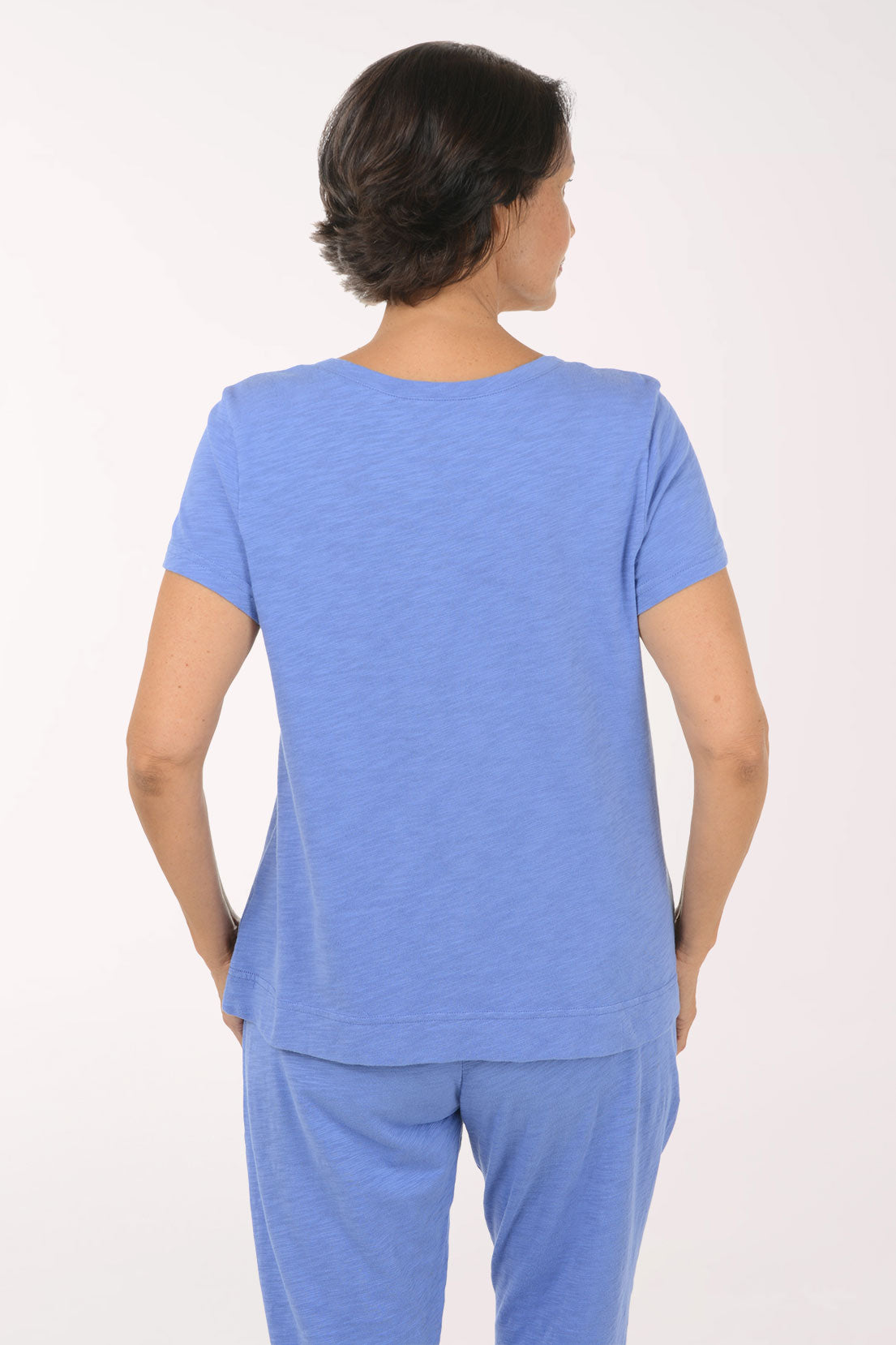 A smiling woman modeling a heathered blue V-neck short-sleeve top with a black dotted heart graphic on the chest, paired with matching blue pants, against a plain white studio background.