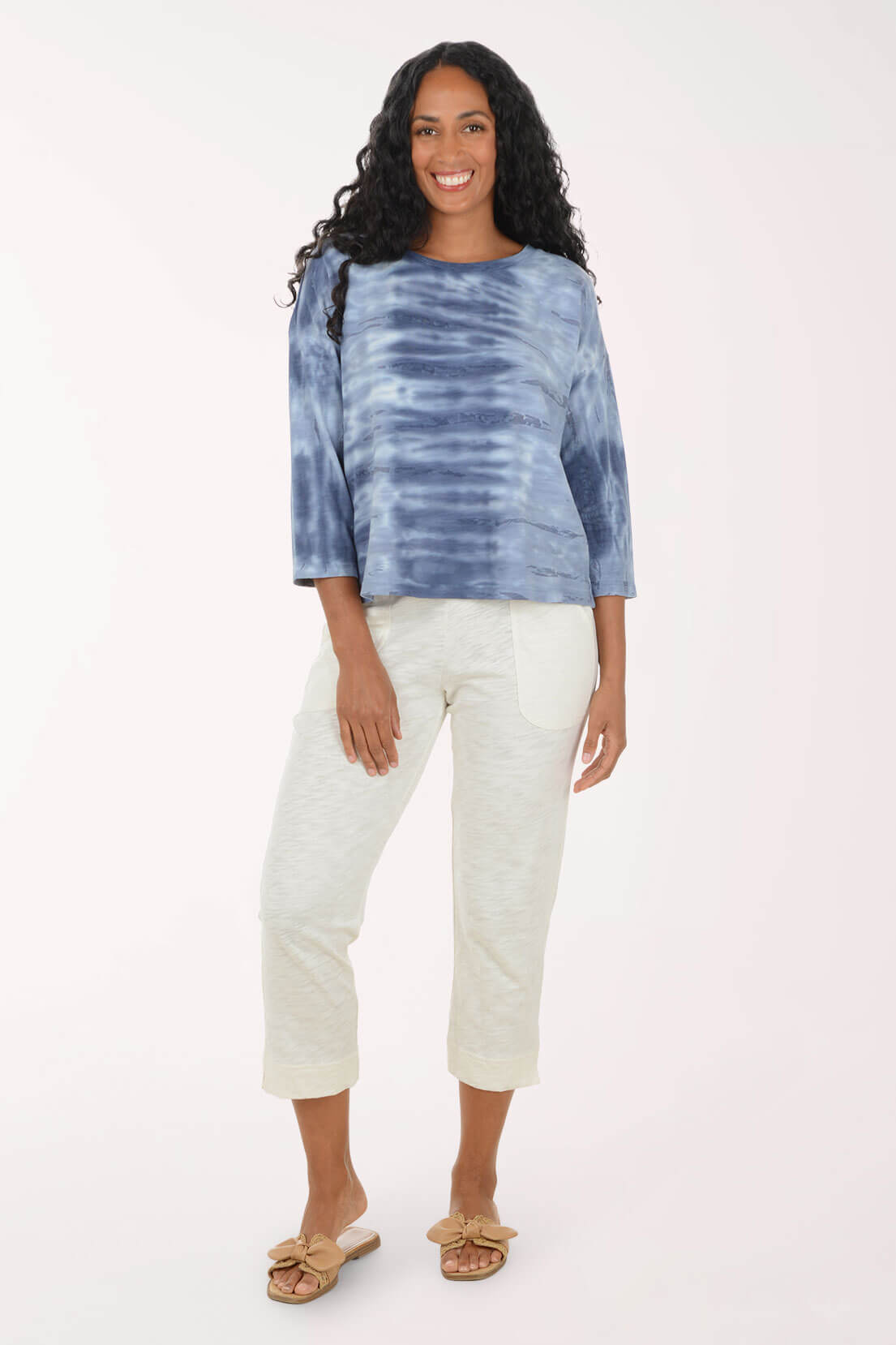 A woman modeling a blue and navy tie-dye long-sleeve top with off-white cropped pants and tan slide sandals, standing against a plain white studio background.