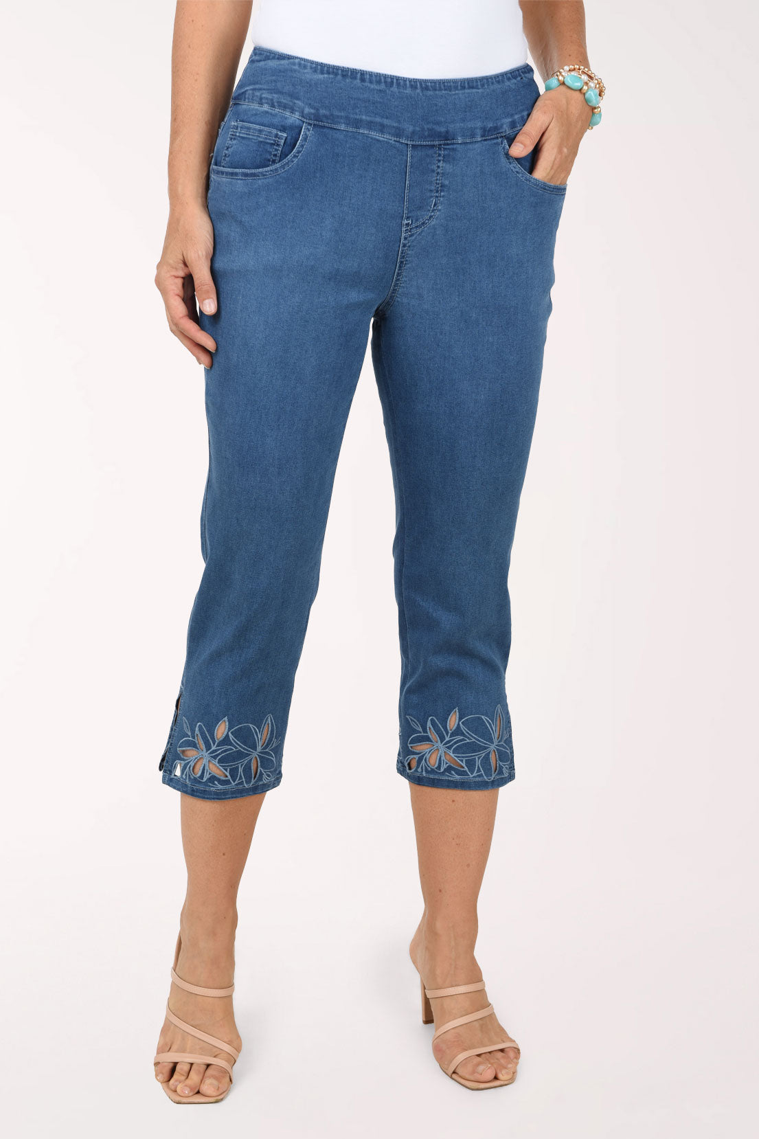A front view of medium blue pull-on denim capri pants with floral cut-out details and side slits at the hem.