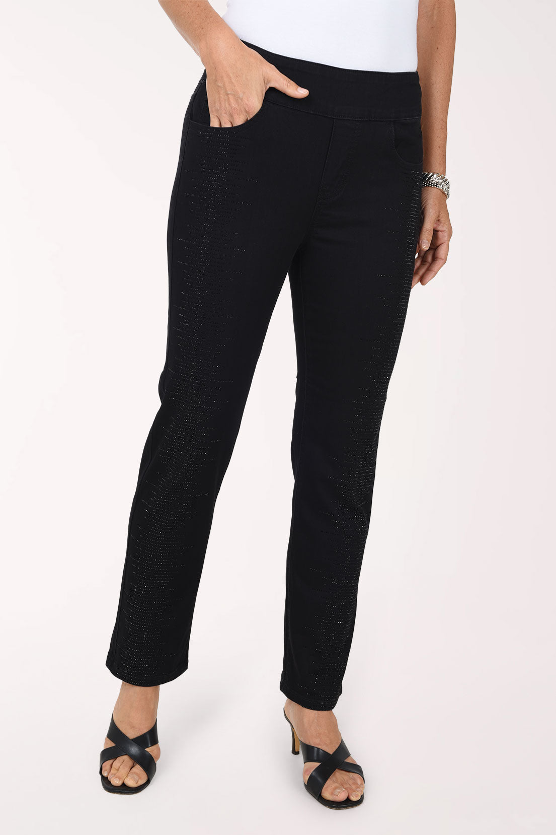 A front view of black straight-leg denim pants with rhinestone embellishment along the legs, worn with a white top and black strappy heels.