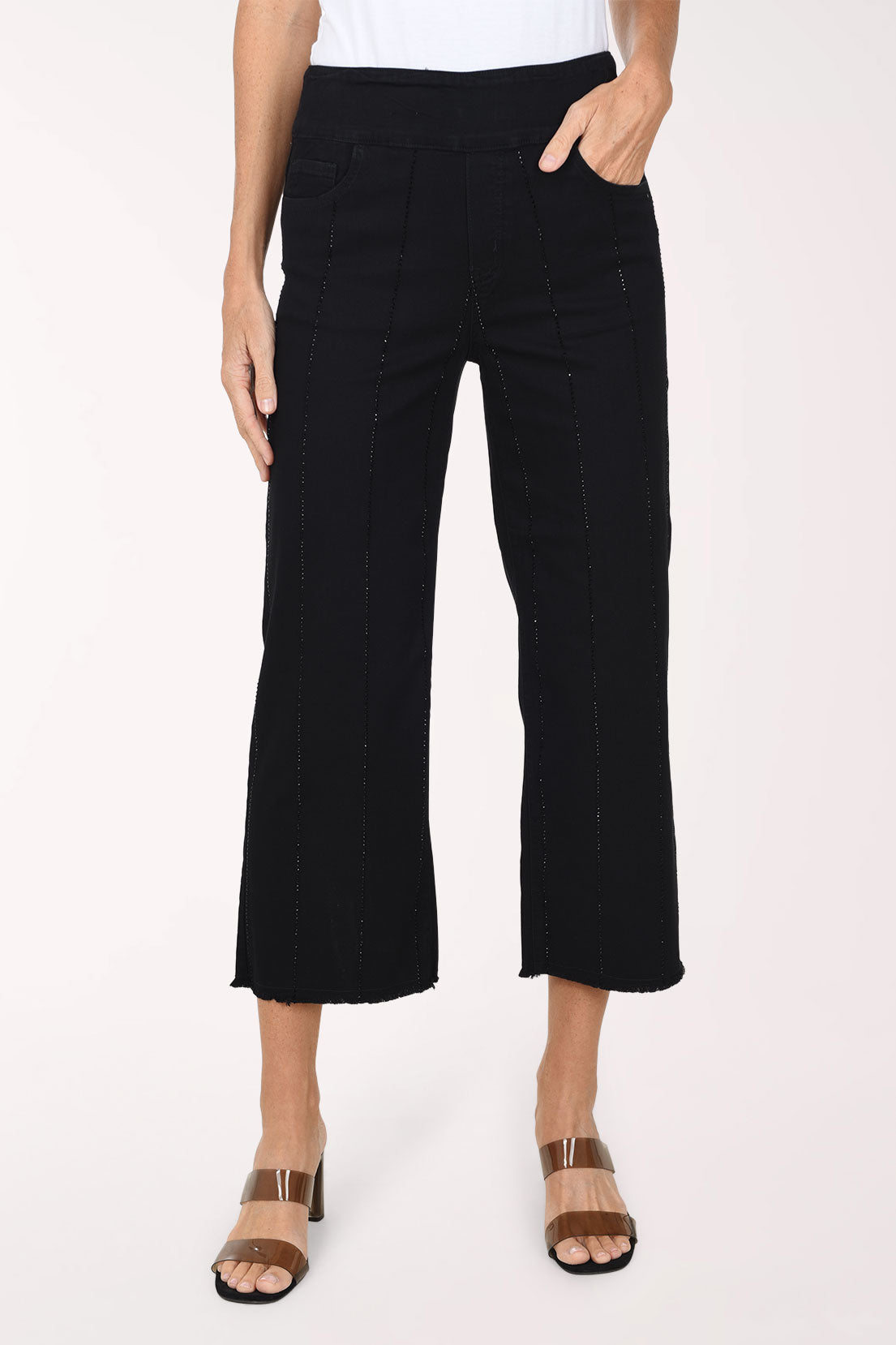 A front view of black wide-leg denim pants with vertical rhinestone stripes, one hand in the pocket, worn with clear-strap heeled sandals.
