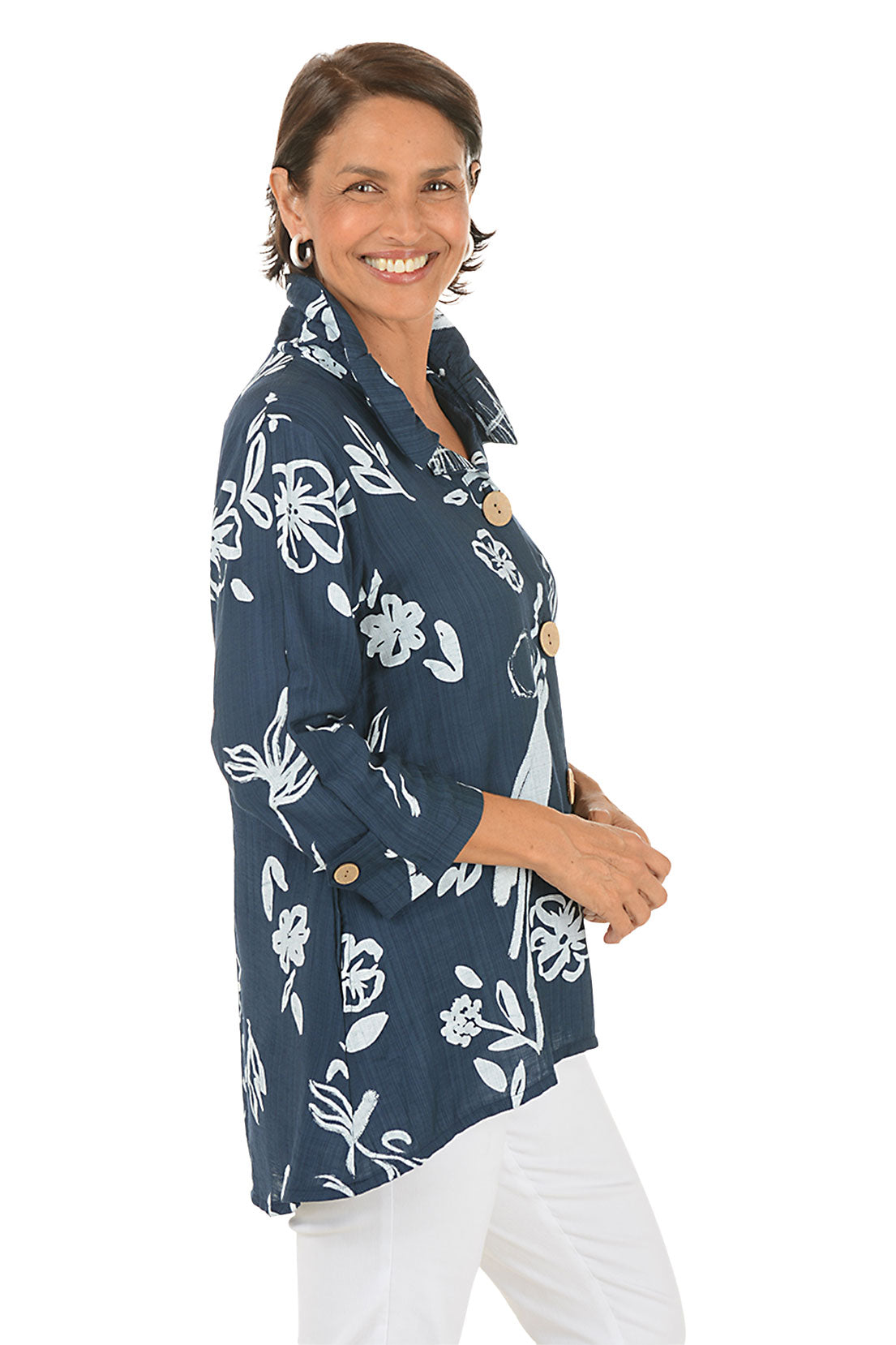 Woman wearing a navy blue floral patterned blouse with white pants on a white background. Side shot.