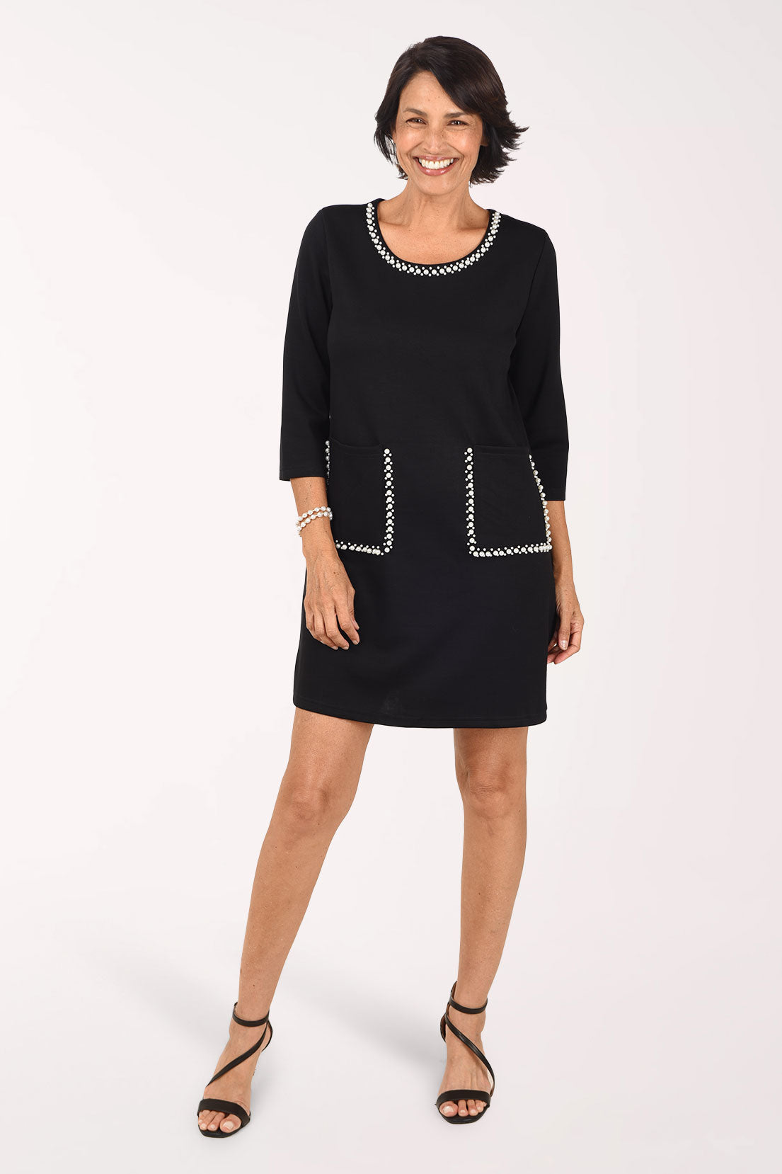 Front view of the black Creation Pearl Trim Pocket Dress, a straight above-the-knee shift with 3/4 sleeves and a scoop neckline edged in white pearl trim, with pearl-trimmed patch pockets at the hips, worn with a pearl bracelet and black strappy heels