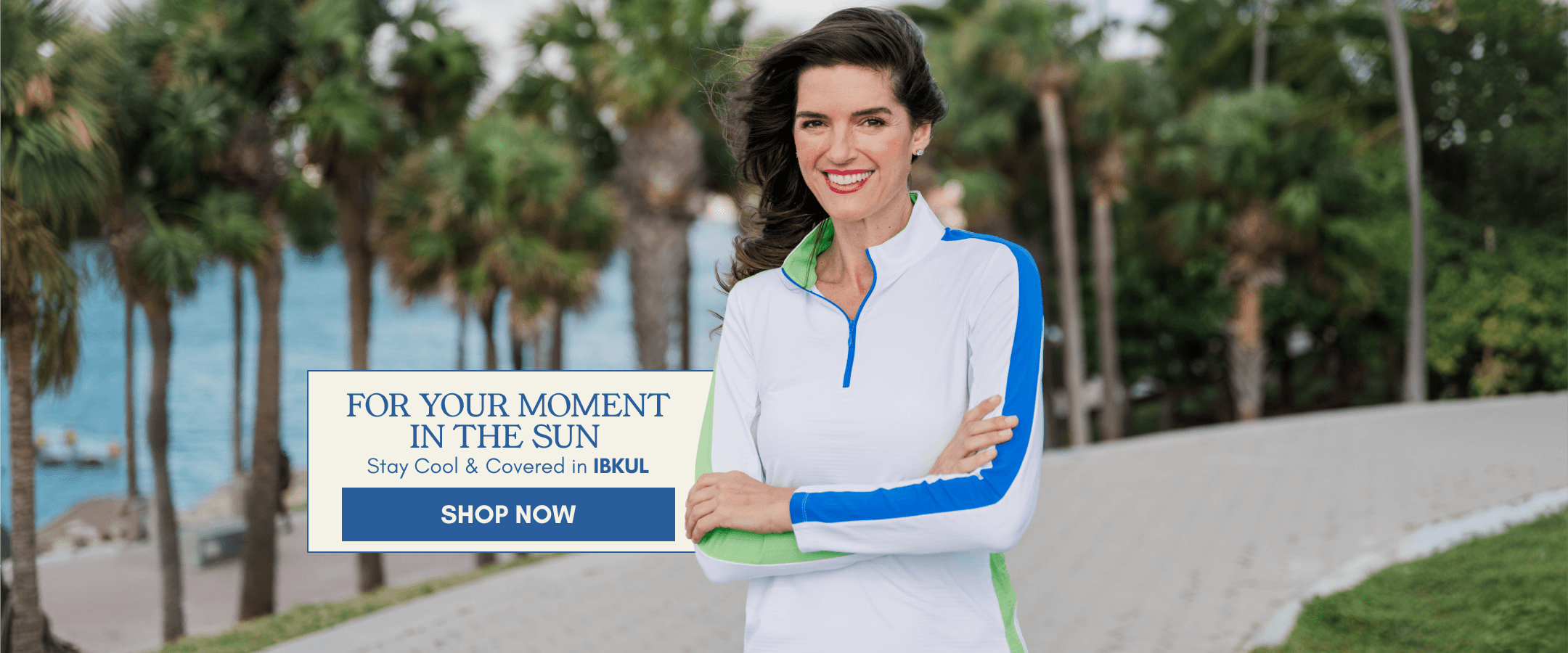 A woman wearing a white athletic zip-up jacket with blue and green trim stands outdoors on a paved path with palm trees and water in the background. A promotional overlay reads 'FOR YOUR MOMENT IN THE SUN Stay Cool & Cooled in IBUL' with a blue 'SHOP NOW' button.
