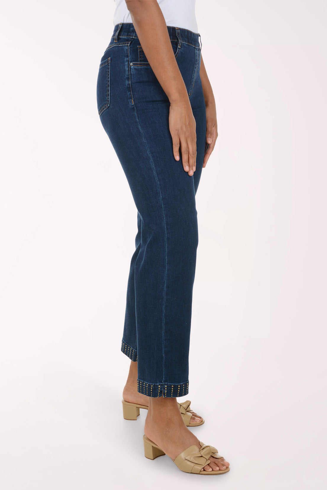 A side-profile view of a model wearing dark blue cropped denim jeans with rolled hems and gold-toned contrast stitching, paired with beige heeled slide sandals against a plain white studio background.