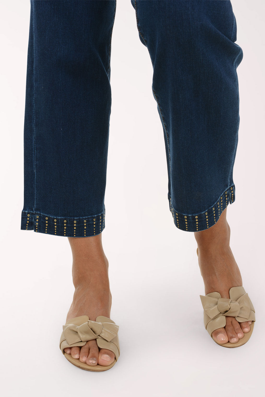 Close-up of a person's lower legs wearing dark blue cropped jeans with decorative gold studs along the hem, paired with beige slide sandals featuring large bow details on the toes, against a plain white background.