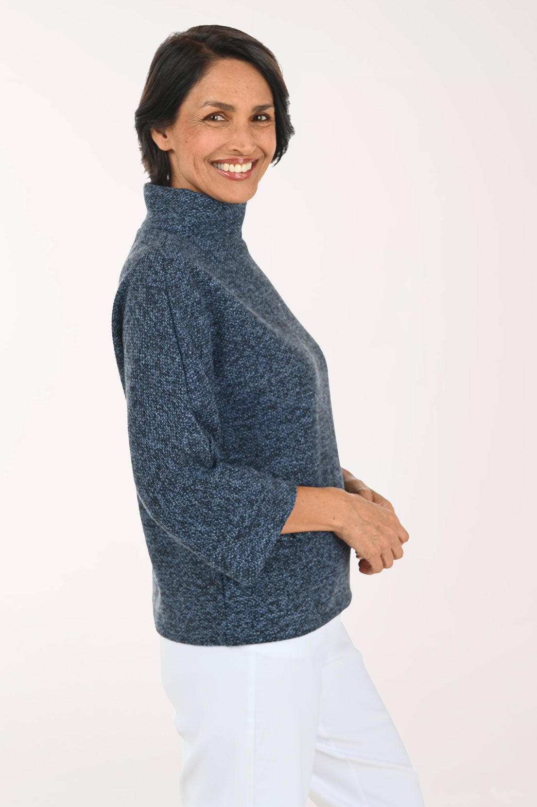 A woman wearing a heathered slate blue mock-neck knit sweater and white pants, photographed against a plain white studio background.