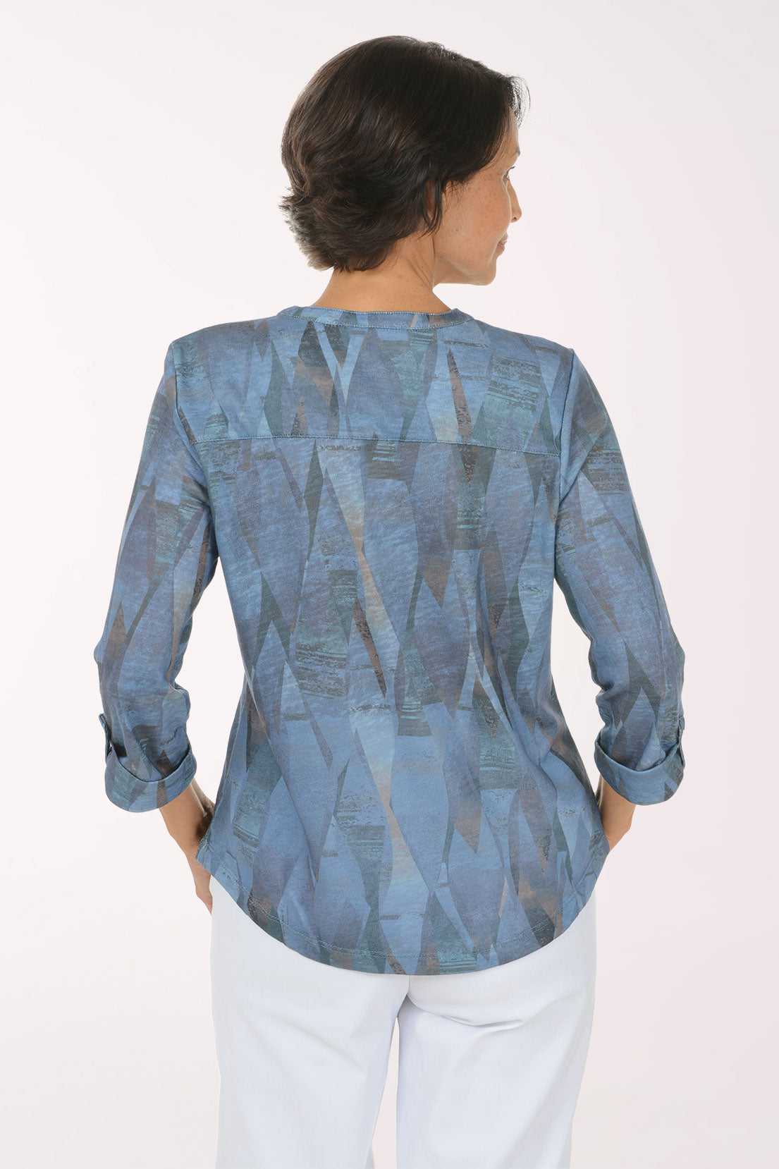 A smiling woman with short dark hair models a blue patterned long-sleeve blouse with white pants against a plain light background.