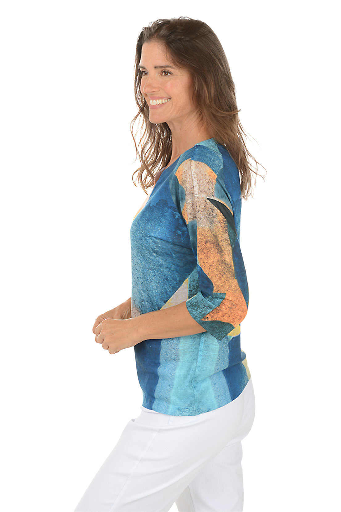 Woman wearing a colorful patterned top with white pants on a white background. Side shot.