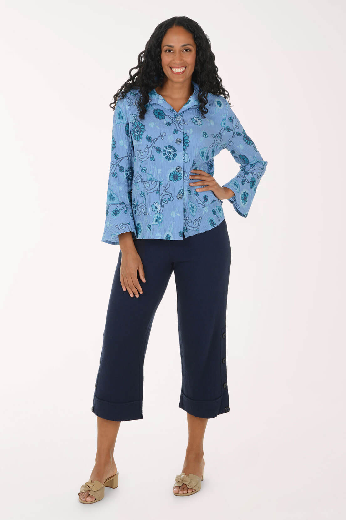 A woman modeling a light blue floral long-sleeve button-up shirt paired with dark navy cropped pants and beige heeled sandals, photographed against a plain white studio background.
