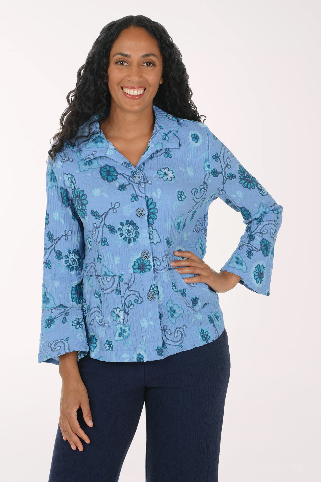 A woman modeling a light blue button-up shirt with a teal and dark blue floral paisley pattern, paired with dark navy pants, against a plain white studio background.