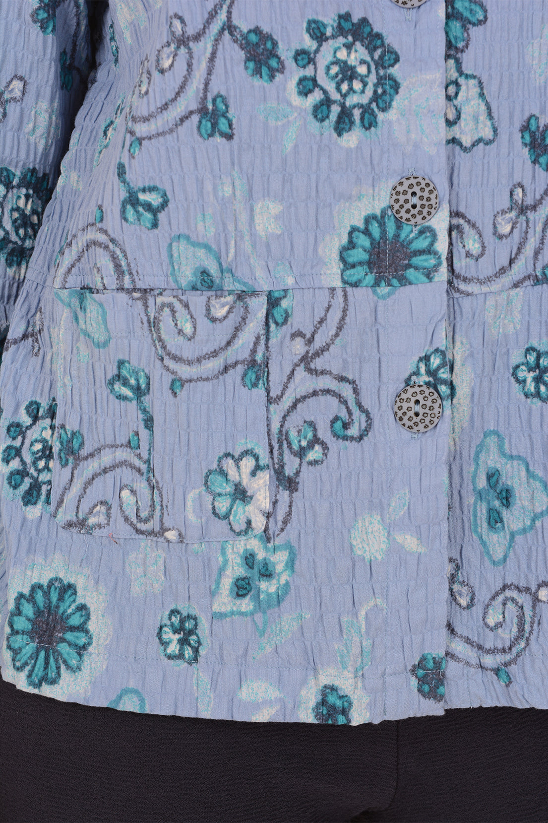 Close-up of a light blue crinkled fabric garment with a teal and white floral pattern, featuring decorative metallic buttons and a pocket seam.