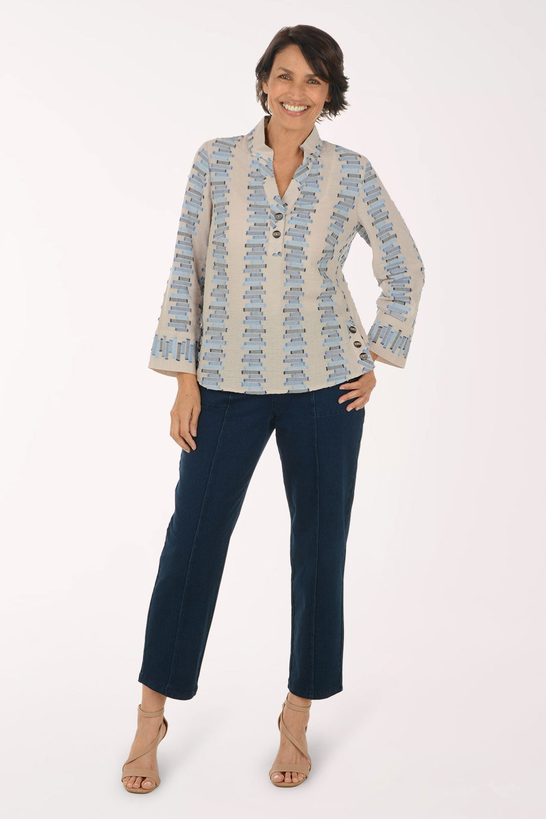 A woman modeling a light beige button-up shirt with a blue and gray geometric pattern, paired with dark navy cropped pants and beige heeled sandals, against a plain white studio background.
