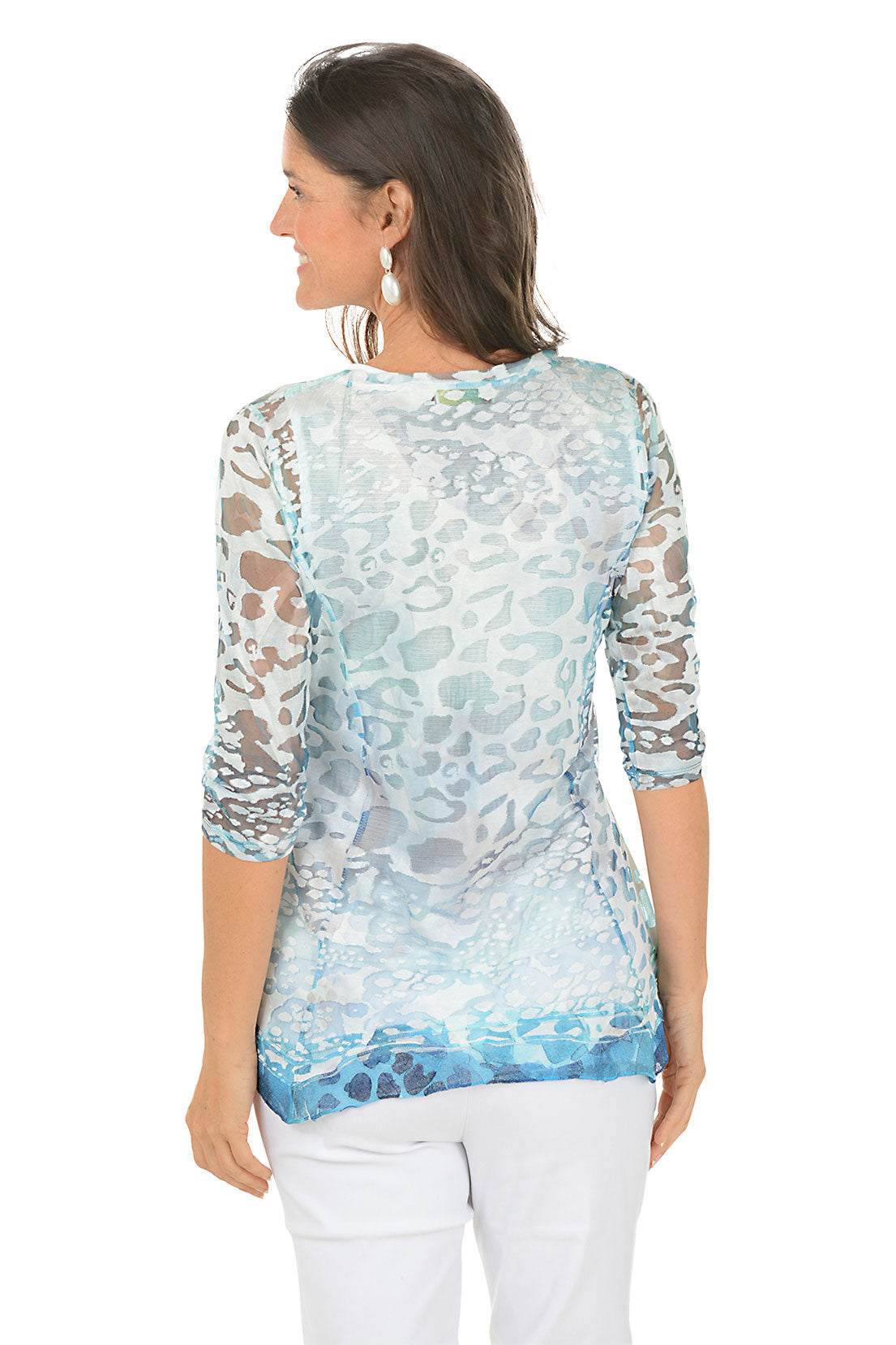 Woman wearing a patterned blue and white blouse with white pants on a white background. Back shot.