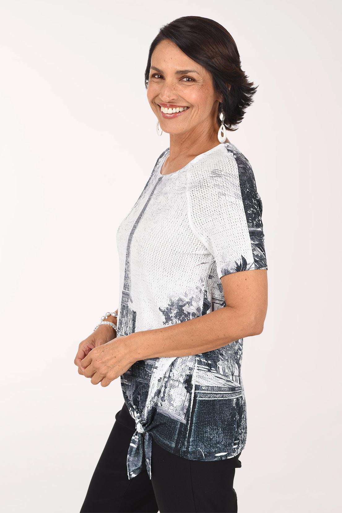 Three-quarter view of the Eiffel Tower eyelet top showing the raglan sleeve and the tie-front hem over black pants.